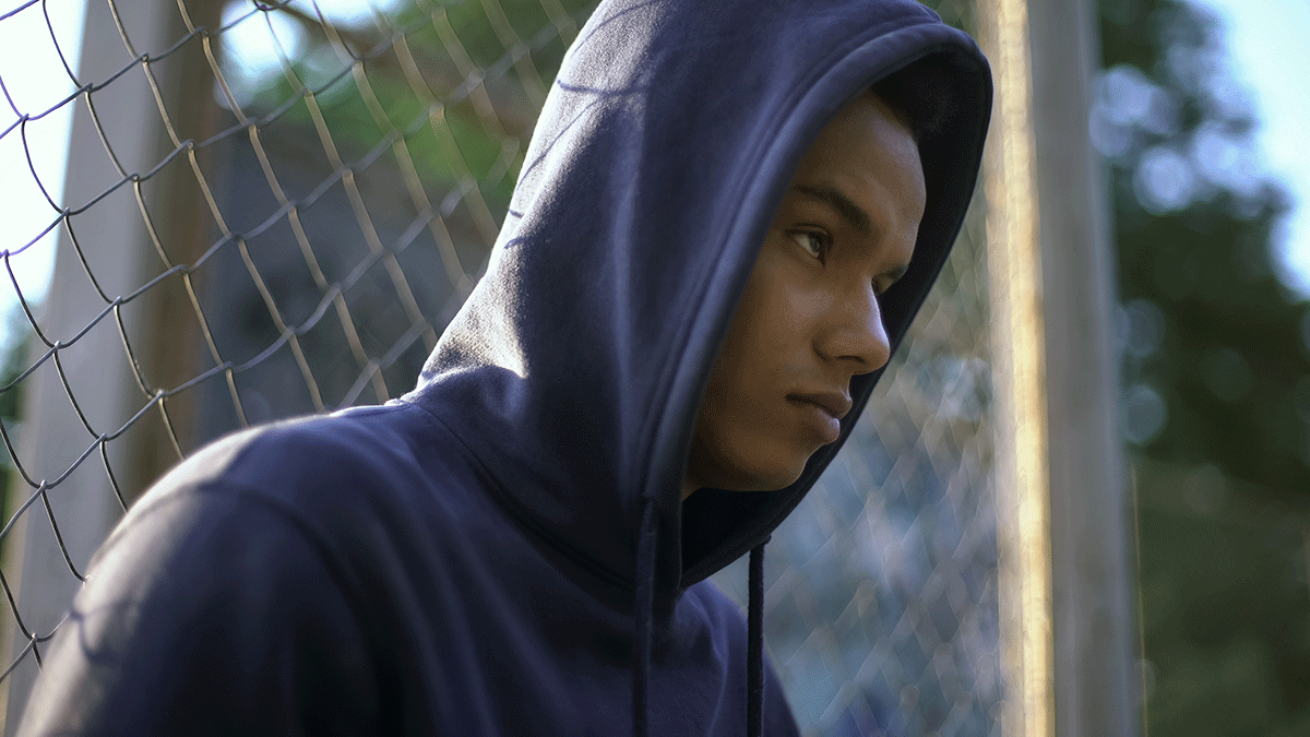 5-Signs-of-Drug-Abuse-In-Teens adolescent wearing hoodie and frowning while sitting against a chain link fence and struggling with signs of drug abuse in teens
