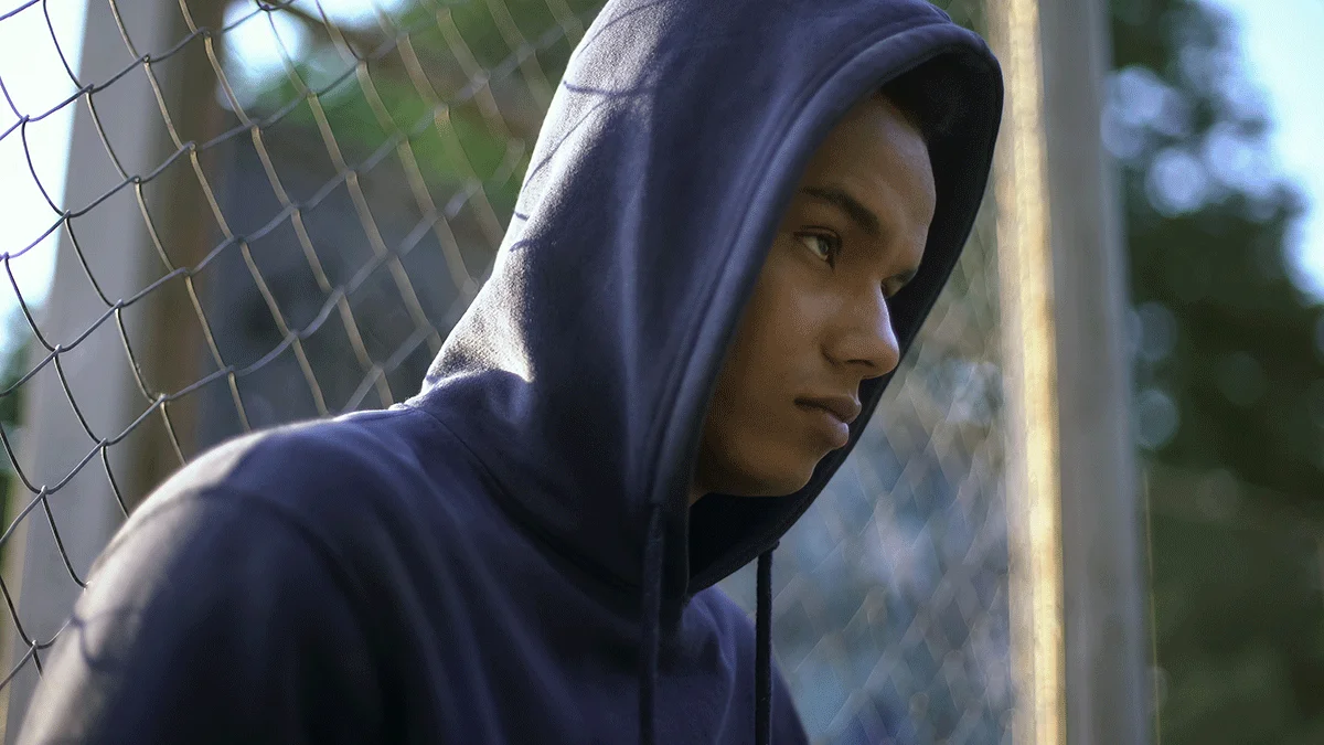 adolescent wearing hoodie and frowning while sitting against a chain link fence and struggling with signs of drug abuse in teens