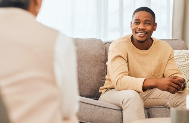 Black man, counseling and psychology consulting for therapy, mental healthcare or support. Happy patient talking to psychologist, therapist and medical help in consultation, advice and wellness check Man smiling while talking with a counselor during a therapy session on a sofa.