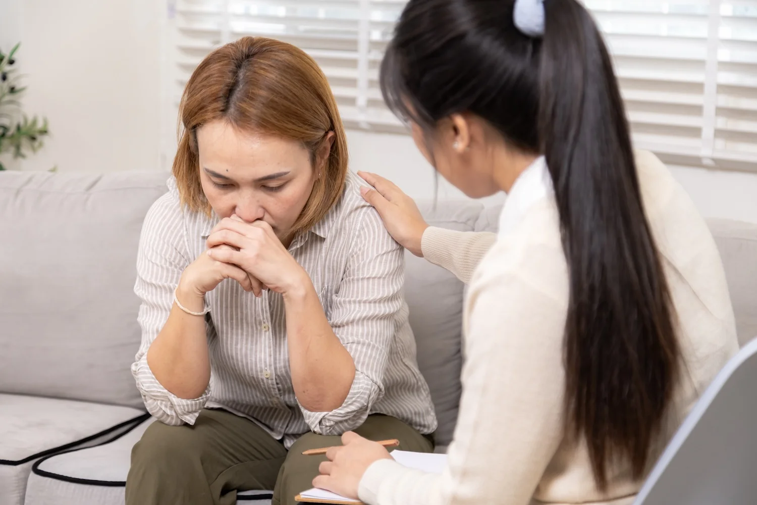 A concerned woman sits on a couch with her hands clasped near her face, while another woman beside her gently places a hand on her shoulder and listens supportively.